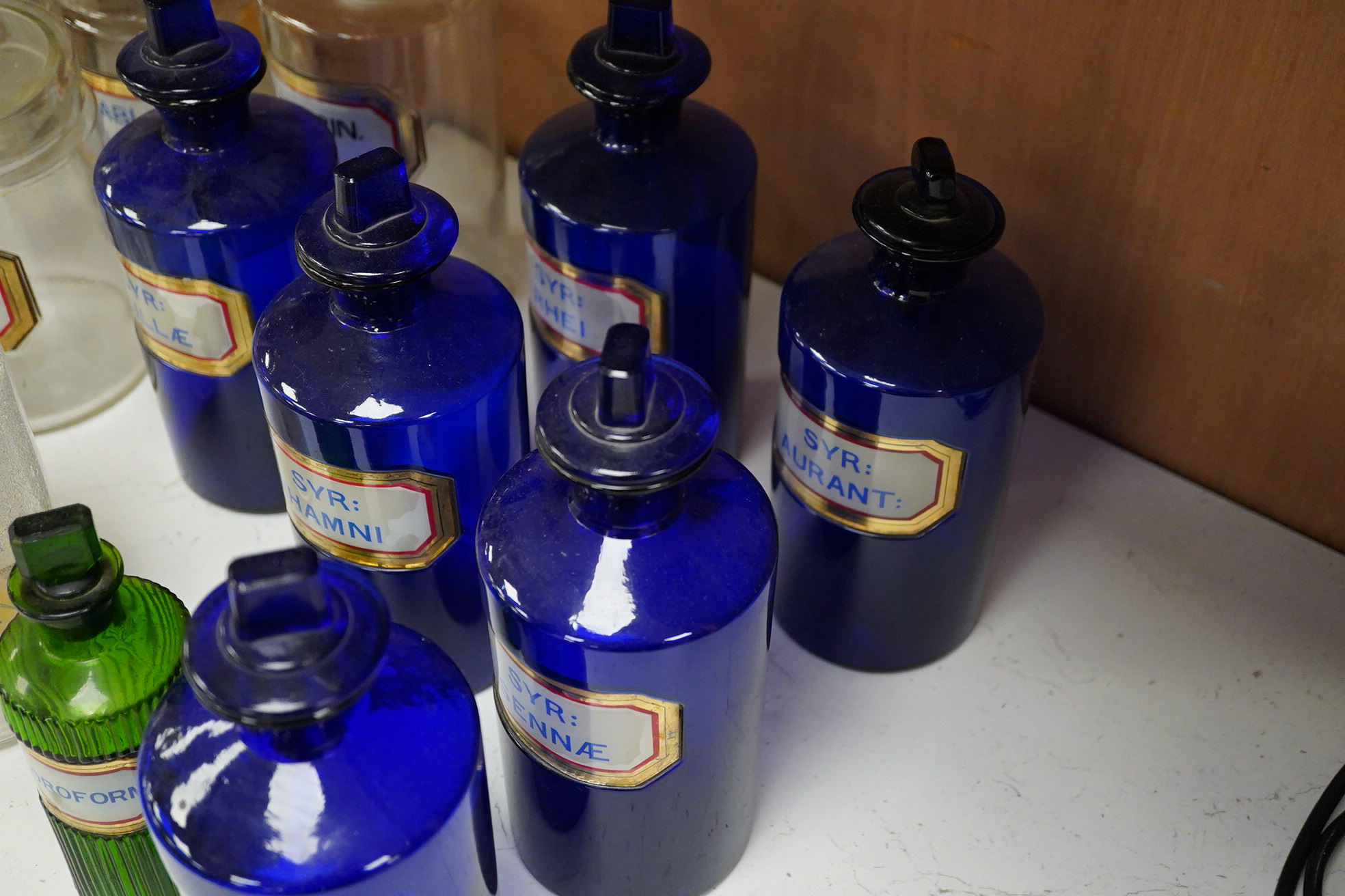 Seventeen 19th/early 20th century glass apothecary jars with gilt rimmed labels; six blue glass, ten clear glass and a green glass example, labels including; Syr: Zingib:, Chloroform, Syr: Rhei:, Camphora, etc. tallest 2